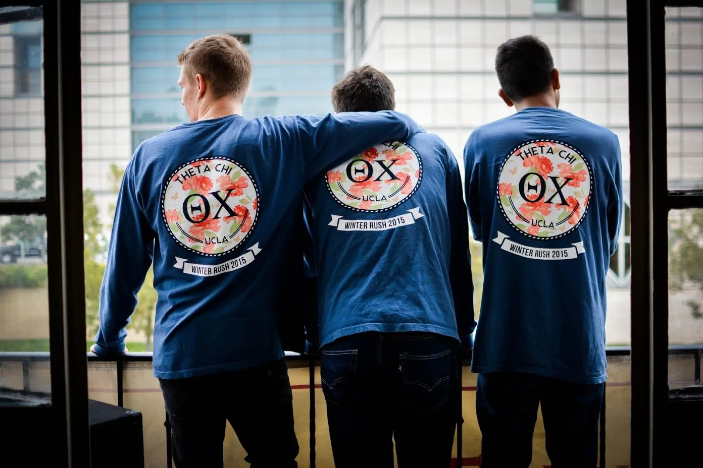 Three brothers in matching Theta Chi rush shirts looking out a window